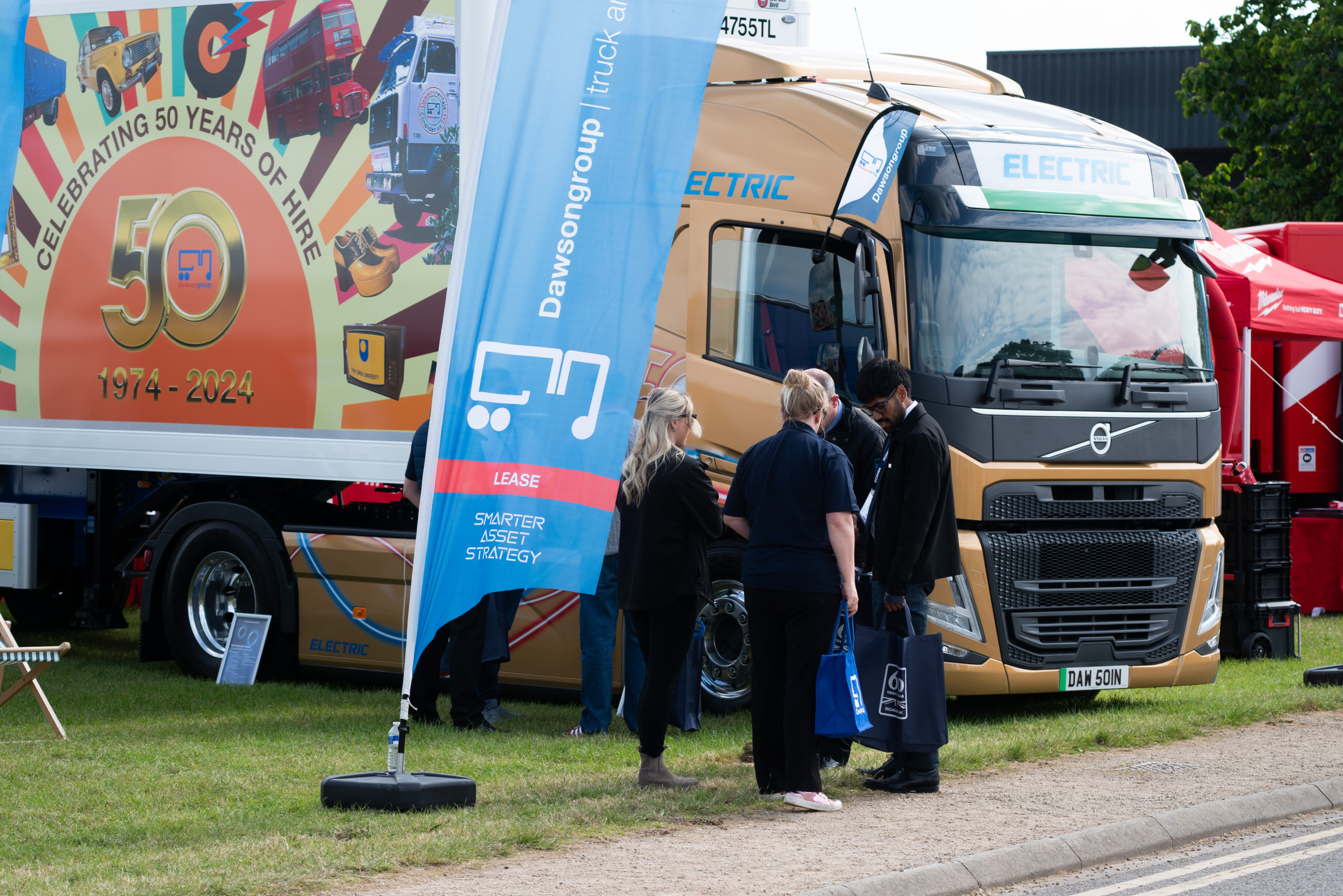 Gold-wrapped Volvo FM Globetrotter 4x2 EV Tractor Unit at the Road Transport Expo 2024. Visitors stand in front, speaking to Dawsongroup truck and trailer employees. In the foreground is a blue feather flag, with lease hire written on it.